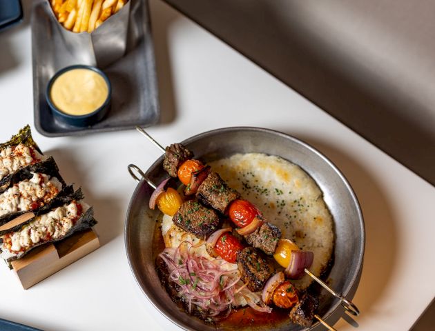 A top-down view of a stylish meal: skewered meat and veggies with a creamy sauce in a metal pan, a side of fries with dipping sauce, and assorted bites on plates.