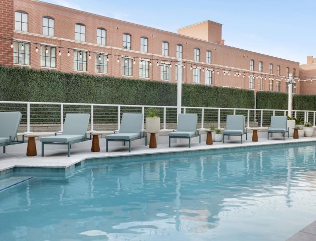 A hotel-style pool area with lounge chairs along the water, a hedge wall, and a brick building in the background.