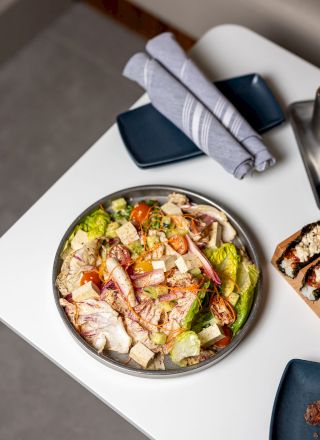 A colorful salad bowl with mixed greens, tomatoes, cucumbers, and grilled chicken pieces on a round plate, with napkins and blue dishes nearby. End sentence.