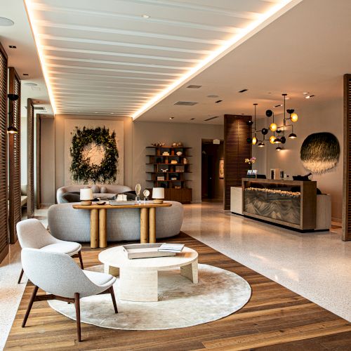 A stylish hotel lobby with modern seating, a round rug, wooden accents, a reception desk, natural light from large windows, and decorative plants, welcoming and bright.