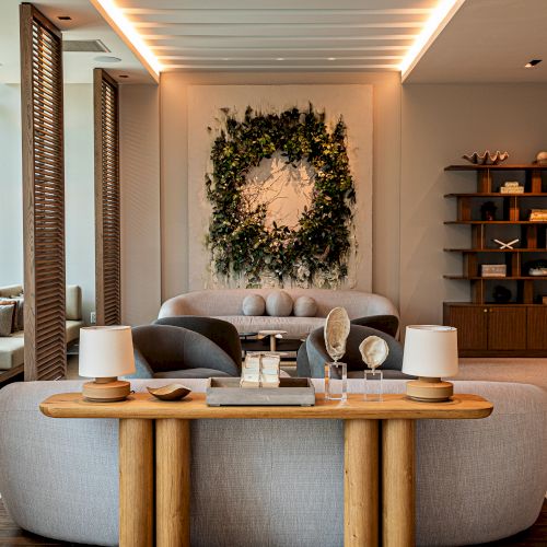 A stylish lobby with a circular gray sofa, a wooden console table, soft lamps, a large green wreath on the wall, and warm ambient lighting.