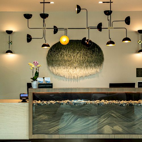 A modern hotel lobby reception with a glass-front desk, a decorative hanging light fixture, and a textured stone feature along the front. End.