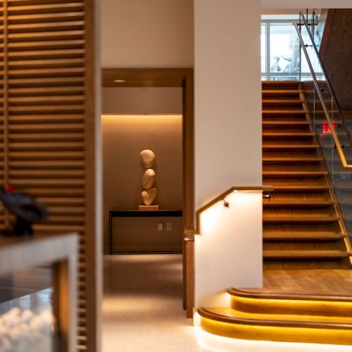 A modern, warmly lit interior with a curved wooden staircase, minimalist decor, and a cozy seating nook near a doorway.