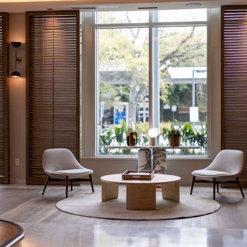 A cozy modern living room with large windows, a low coffee table, plants, and natural light streaming in through the doorway.
