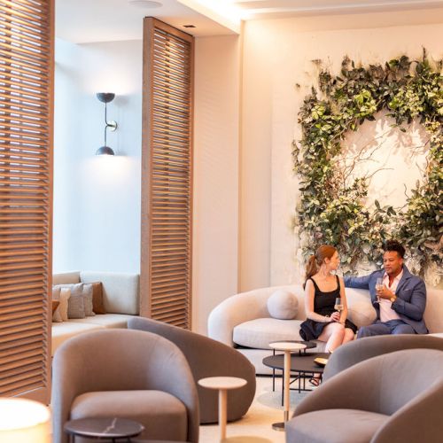 A cozy living room with a wicker chair, a patterned rug, a wreath on the wall, and warm natural light from a window.