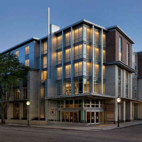 A modern multi-story building with brick and glass windows, warmly lit interiors, and a street-level entrance on a clear evening.