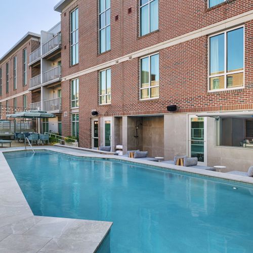 A modern brick building&rsquo;s courtyard patio with a blue swimming pool, lounge chairs, string lights, and large windows.