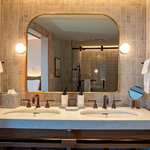 A bathroom vanity with a double sink, large arched mirror, wall sconces, tiled walls, towels, and organized toiletries, all neatly arranged.