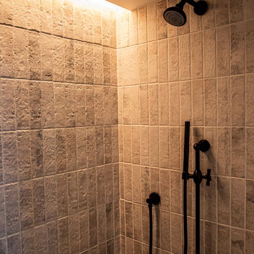A tiled shower with a black adjustable handheld shower bar and a ceiling-mounted rainfall showerhead, lit by warm ceiling lighting.