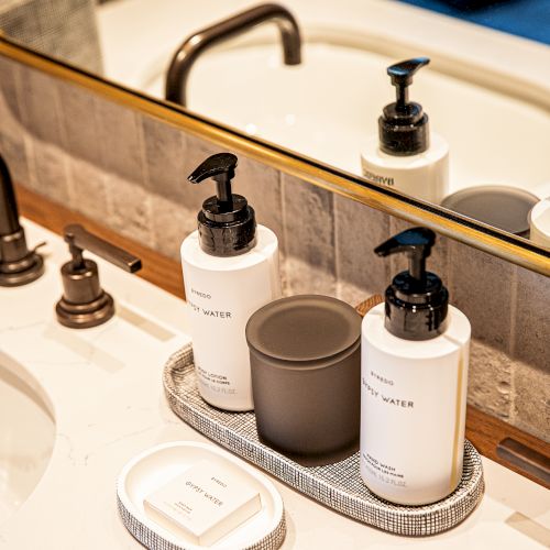 Toiletries on a bathroom sink: white bottle dispensers with pumps, a small round gray holder, soap bar, and a silver tray, mirrors reflect the setup.