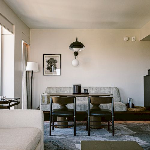 A modern living area with a gray sofa, a small dining table and four chairs, a bar cart by tall windows, and minimalist decor including a floor lamp and wall art.