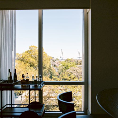A hotel room with a large window view of trees outside, a desk with bottles and glasses, and a curved chair facing the window, bright daytime light.