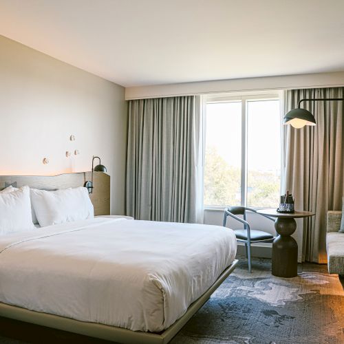 A modern hotel room with a large bed, two lamps, a sitting area by a window, soft gray curtains, and neutral decor. End with a period.