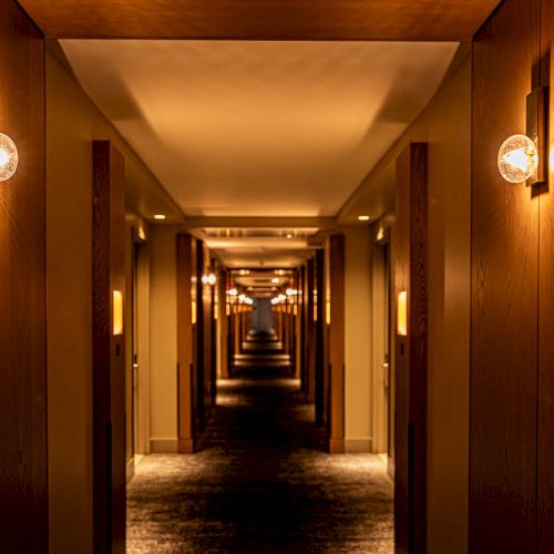 A long, dimly lit hotel corridor with warm wall lamps and a repeating perspective that vanishes into the distance.