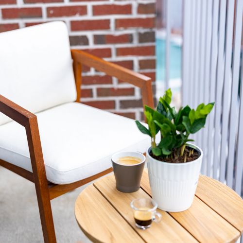 A small outdoor seating area with a wooden table, two chairs, a white mug with a plant, and a brick wall backdrop.