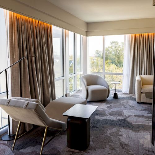 A bright hotel room with a window view, modern decor, a bed, seating area, and soft natural light.
