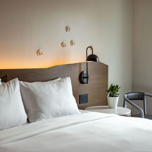 A tidy hotel bedroom with a low wooden headboard, white bedding, two pillows, and a small bedside table with a lamp.