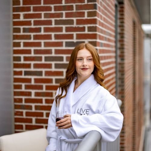 A smiling woman sits on stairs against a brick wall, wearing a white top and light cardigan, posing casually with a striped blanket or sign.