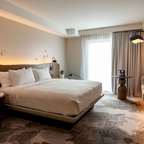 A cozy hotel bedroom with a large bed, warm lighting, a seating area, and a bright windowed corner. Top it at 140 characters, always ending the sentence.