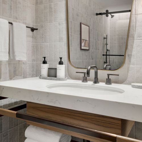 A modern bathroom with a white marble sink, chrome faucets, towels, soap dispensers, and a large illuminated mirror with shelf-like d&eacute;cor.