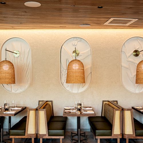 A row of four people sitting in chairs, each with a round arch framed backdrop behind them, in a bright, hallway-like setting. End the sentence.