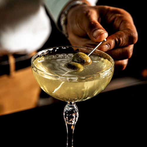 A bartender stirs a classic cocktail in a coupe glass, garnished with an olive on a pick, set against a dim, warm bar backdrop.