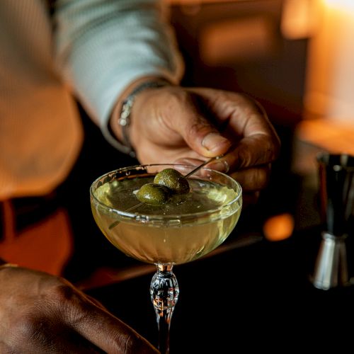 A bartender pours a cocktail into a glass as a hand decorates it; warm, dim bar lighting in the background.