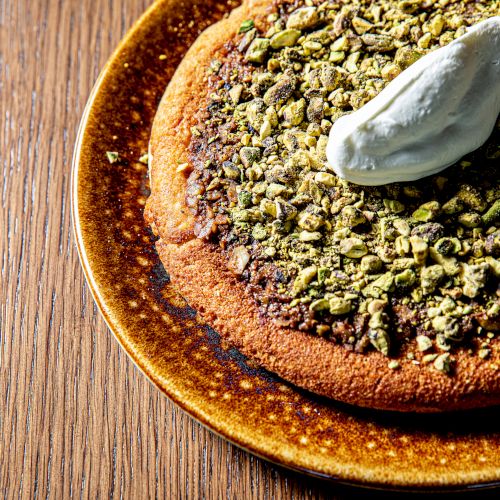 A small round plate with a handful of greenish seeds or grains on a wooden surface, presented sparsely, plus a single green herb leaf.
