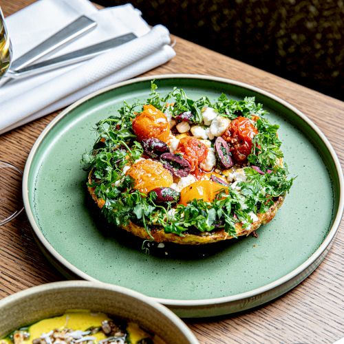 A bowl of green salad on top of white rice with colorful toppings, citrus wedges, and a wooden plate nearby.