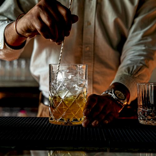 A chef with a tall hat flamb&eacute;ing a pan over a blazing stove in a busy, rustic kitchen.