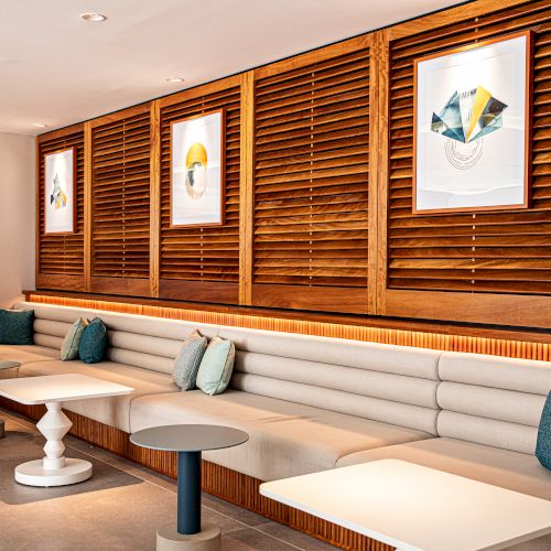 A modern lounge with a long white sofa, blue cushions, wooden slat walls, round white tables, and framed art posters along the wall.