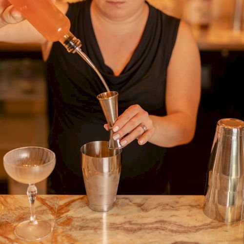 A person is pouring a drink into a glass on a table, with hands near a bottle and other glassware; warm lighting and a casual setting.