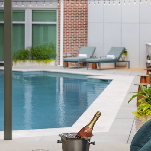 A luxury patio with a swimming pool, lounge chairs, and a chilled champagne bottle in an ice bucket by the edge.