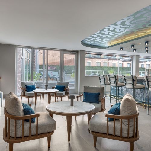 Modern lounge with cushioned chairs, round coffee tables, large windows, and a patterned ceiling mural.
