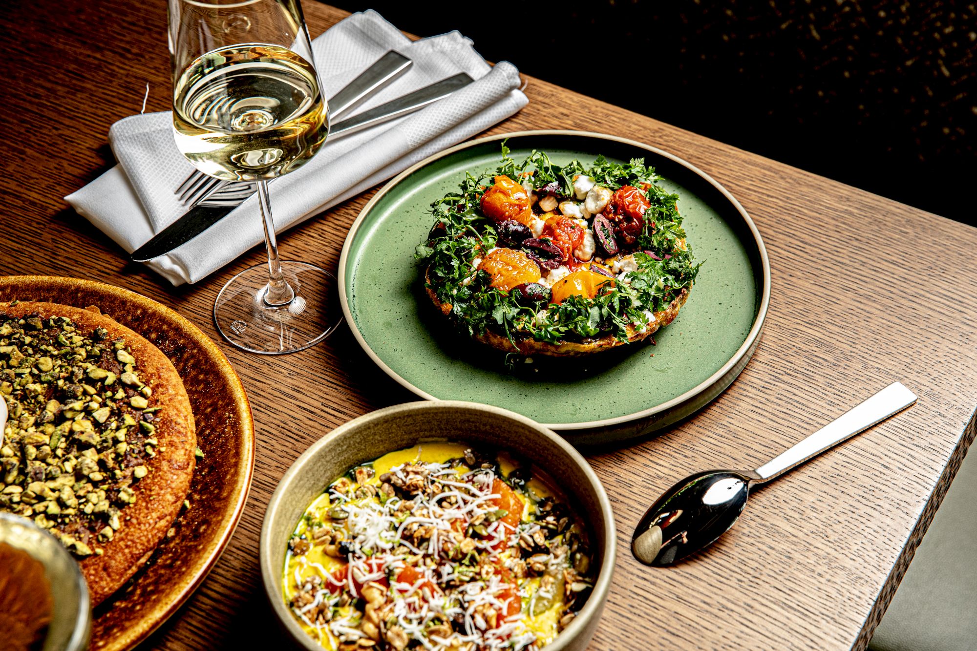 A plated kale salad on a green plate, creamy bowl with grains, a slice of quiche or tart nearby, a glass of white wine, and a spoon on a wooden table.