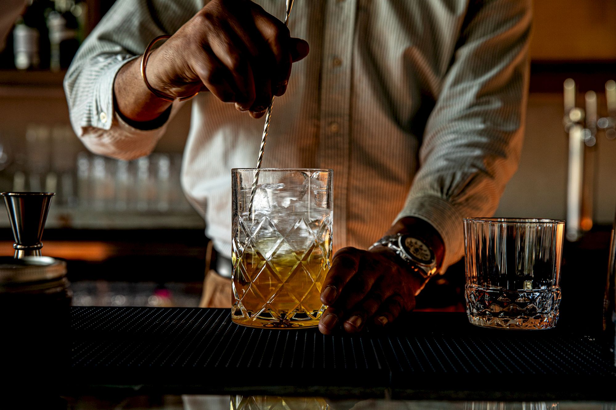 A bartender stirs a whiskey sour over ice in a glass, with a second glass beside it on the bar.