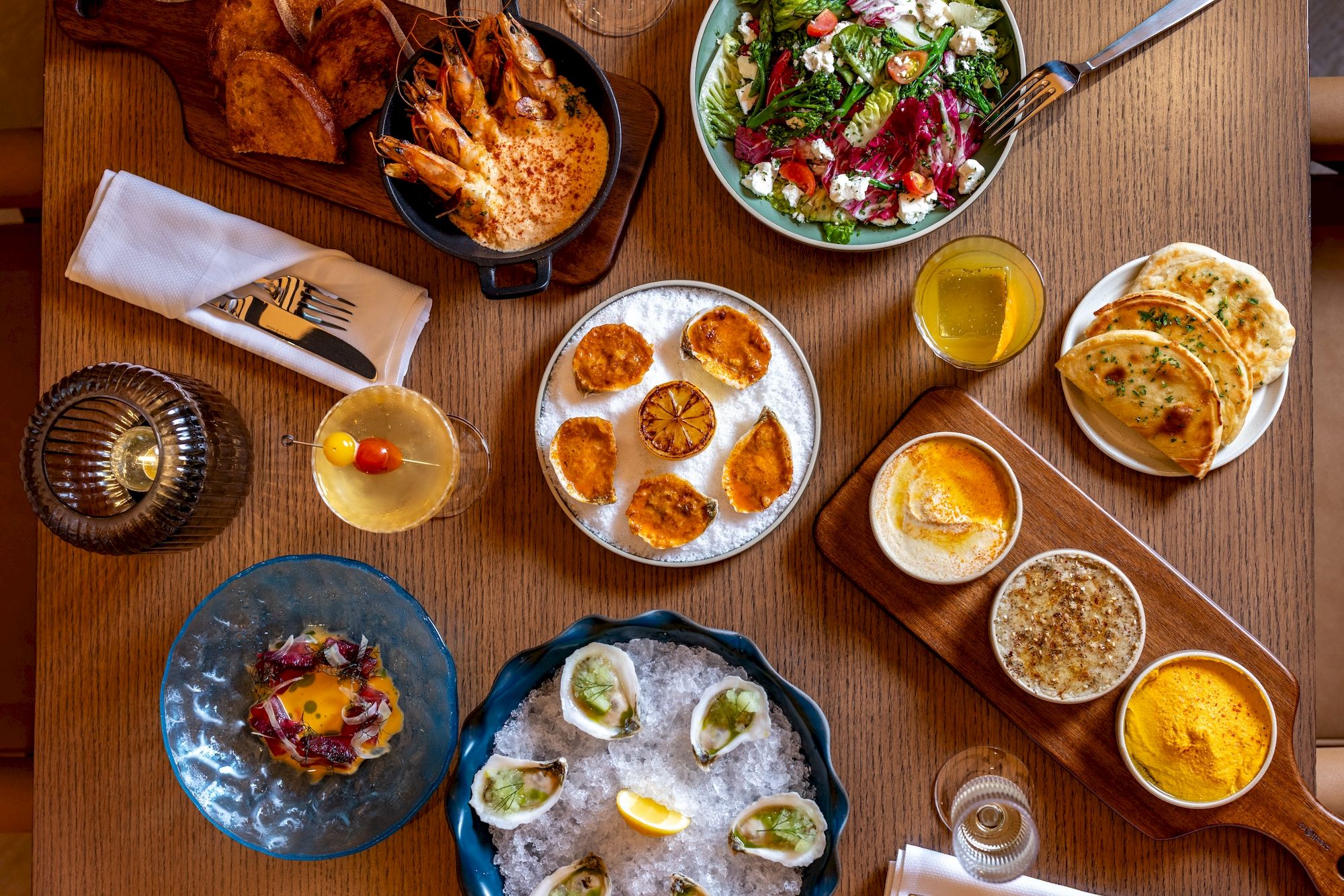 A top-view spread of plates and bowls with breakfast items: pancakes, dumplings, scones, eggs, salad, croissants, jam, coffee, tea, and juices.