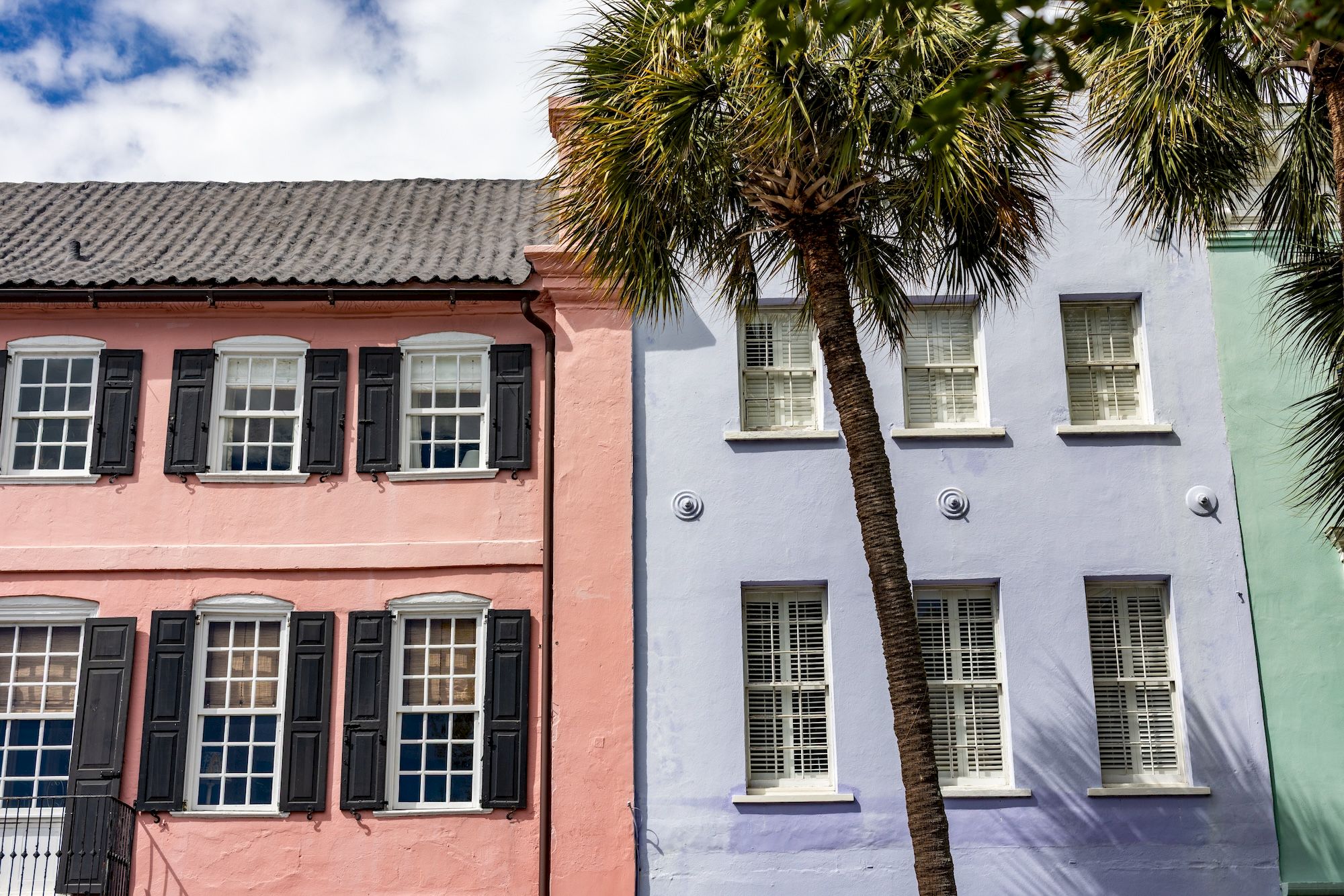 Colorful row houses with pastel pink, lavender, and teal facades, tall palm trees leaning between buildings, bright windows, sunny day.