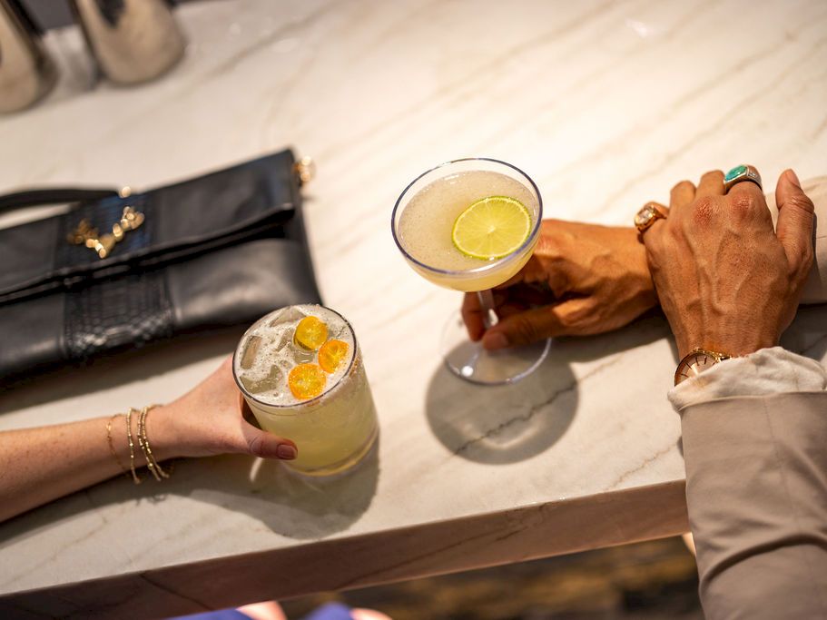 Two people at a bar clink cocktails: one with a lemon slice, the other with a lime slice, on a marble counter with a dark purse nearby.