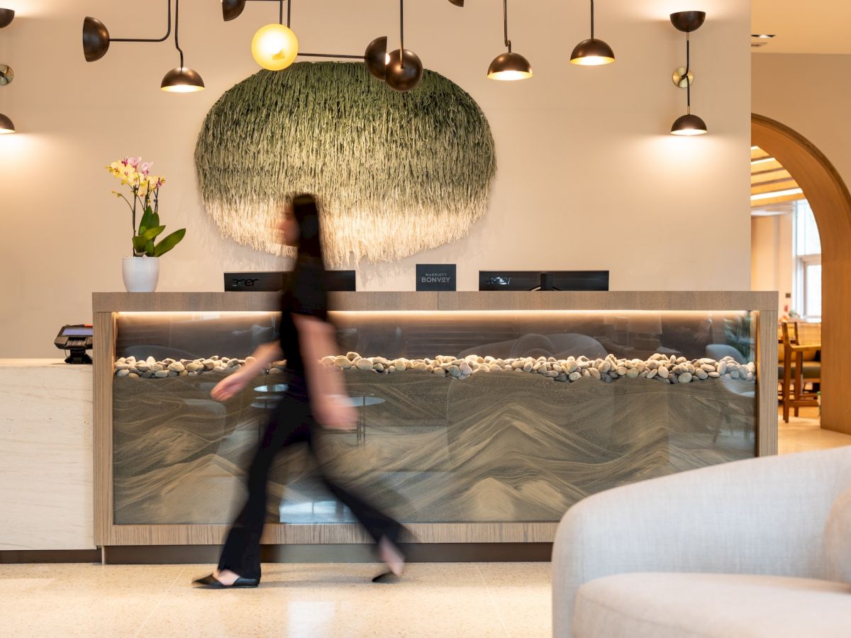 A blurred person walks past a modern reception desk with a wave-pattern panel, pendant lights, and a green hanging art piece above the counter, in a hotel lobby.