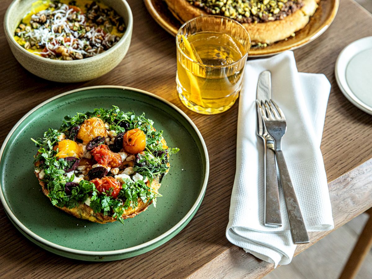 Fresh salad with greens, tomatoes, nuts on toast, plus a beer and a pizza in the background; cozy cafe vibes.