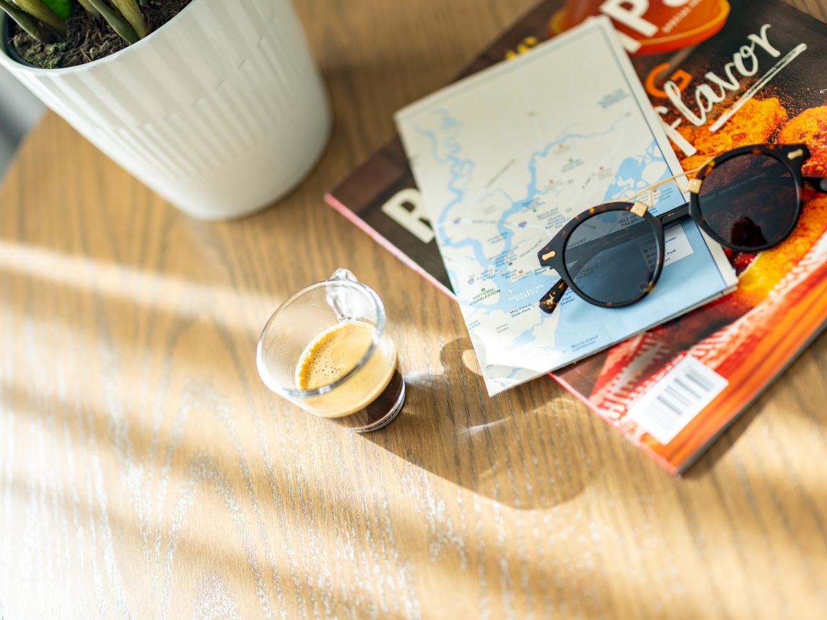 A round wooden table with a plant, sunglasses, a postcard, a small cup of espresso, and a couple of magazines stacked beside sunlight stripes streaming across the surface.