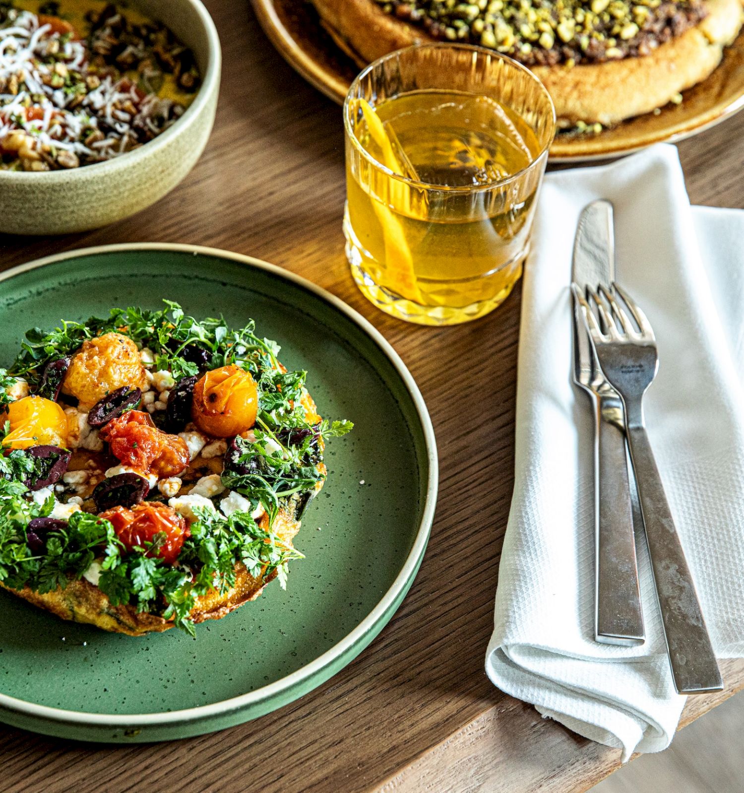A fresh salad on a green plate topped with greens, tomatoes, and orange chunks; beside it are a beer, a fork, knife, and napkin on a wooden table.