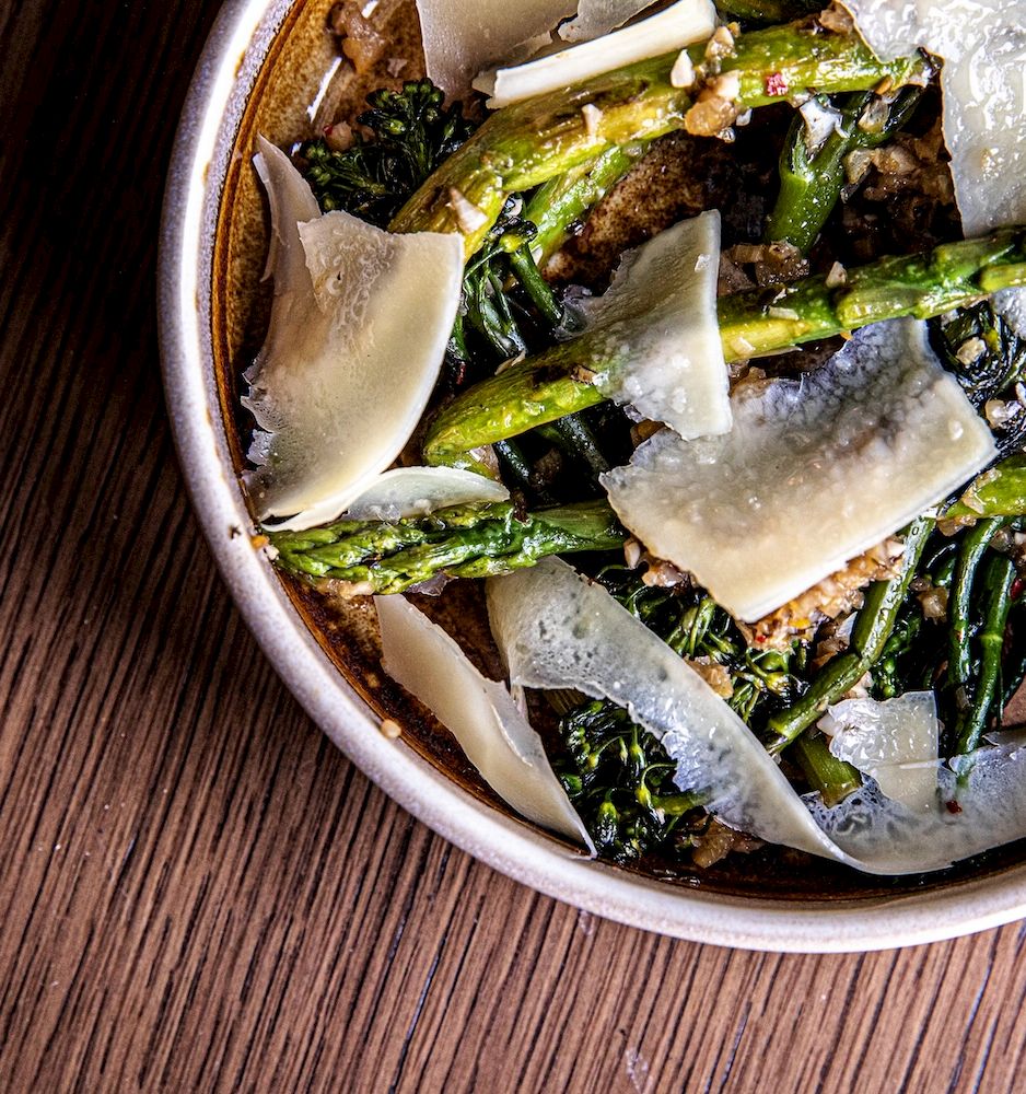 A bowl of roasted asparagus with shavings of cheese on a wooden table, torn crisp bits mixed in.
