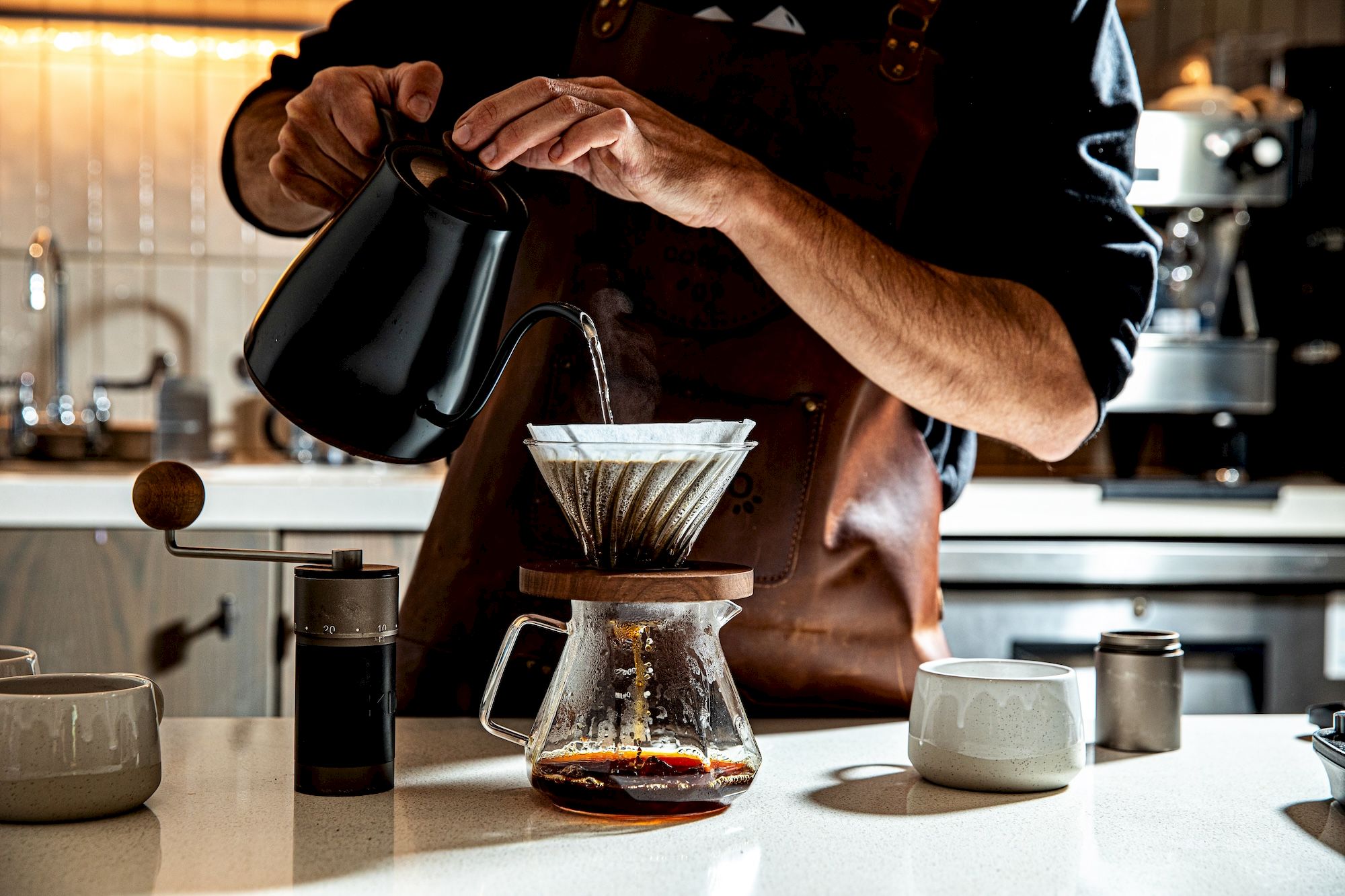A barista pours hot water through a coffee filter into a Chemex, brewing coffee on a white countertop with a grinder and kettle nearby.