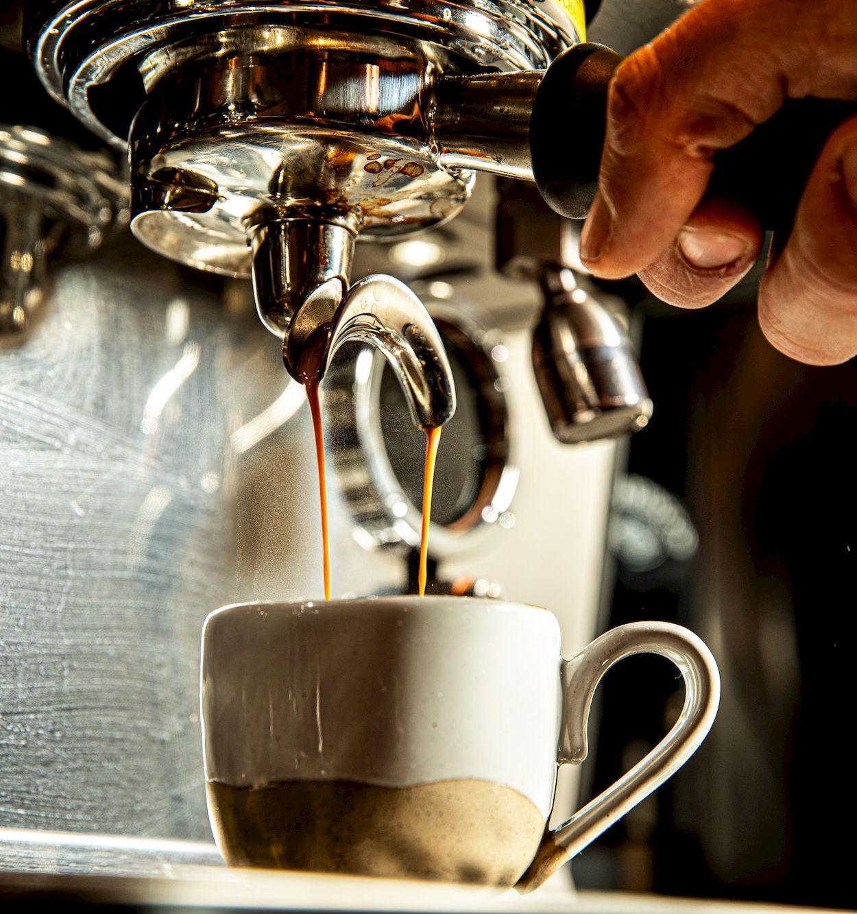 A coffee machine pouring espresso into a small white cup, steam rising as the spout drips coffee, held by a barista&rsquo;s hand.