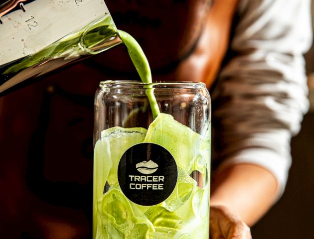 A barista pours vibrant green liquid into a glass with ice, from a jug marked &ldquo;Tracer Coffee,&rdquo; creating a fresh, layered drink.