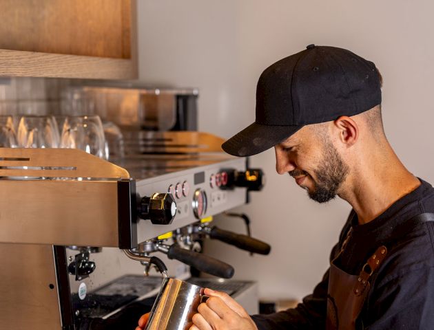A barista wearing a cap and apron operates a professional espresso machine, steaming milk into a metal pitcher.