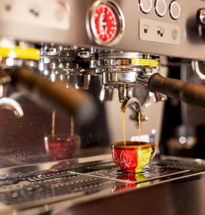 A shiny espresso machine pours a two-toned shot into a small red-and-yellow demitasse cup, steam wand at the ready, timer blinking.