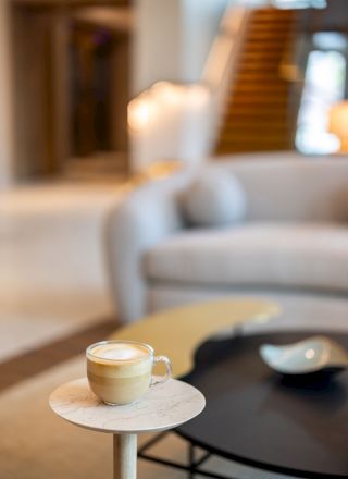 A cozy living room with a latte on a tiny marble side table, soft seating in the background, and a warm, blurred staircase in view.